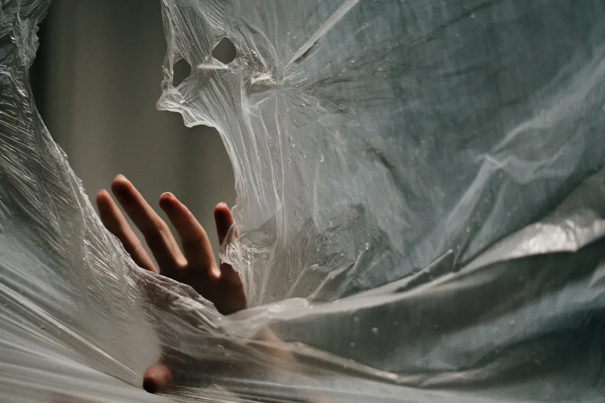 A hand pushing through thin, crumpled plastic, suggesting themes of entrapment and freedom.