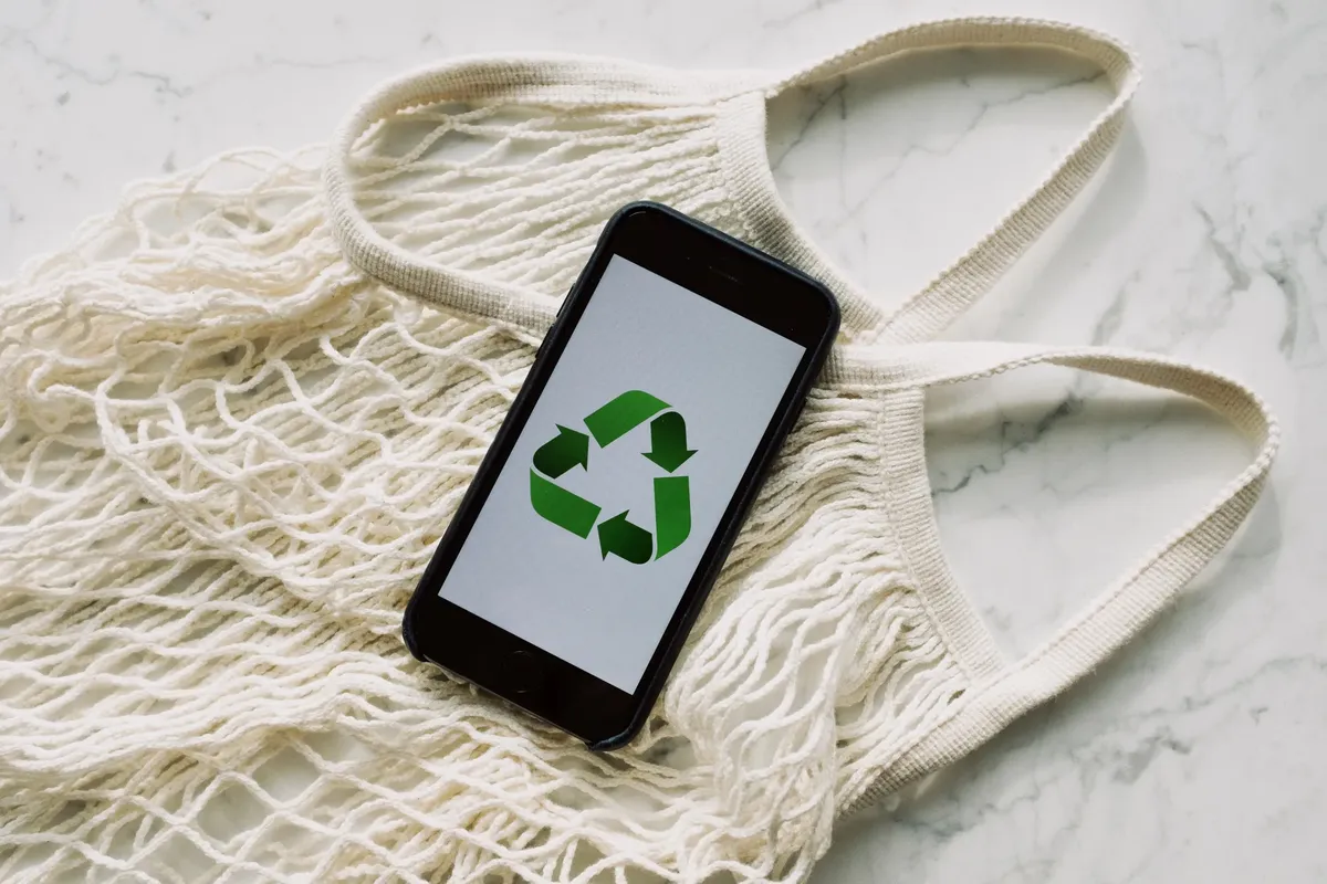 Top view of mobile phone with green recycling emblem on screen composed on white mesh bag on marble table in room